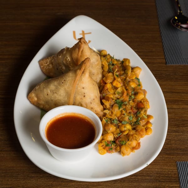 Two golden-brown samosas, a side of chickpea curry, and a small bowl of red dipping sauce on a white plate.