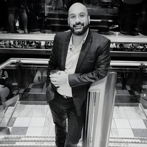 A smiling man in a suit and glasses holds a drink, leaning on a railing in a restaurant.