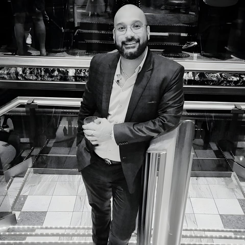 A smiling man in a suit and glasses holds a drink, leaning on a railing in a restaurant.