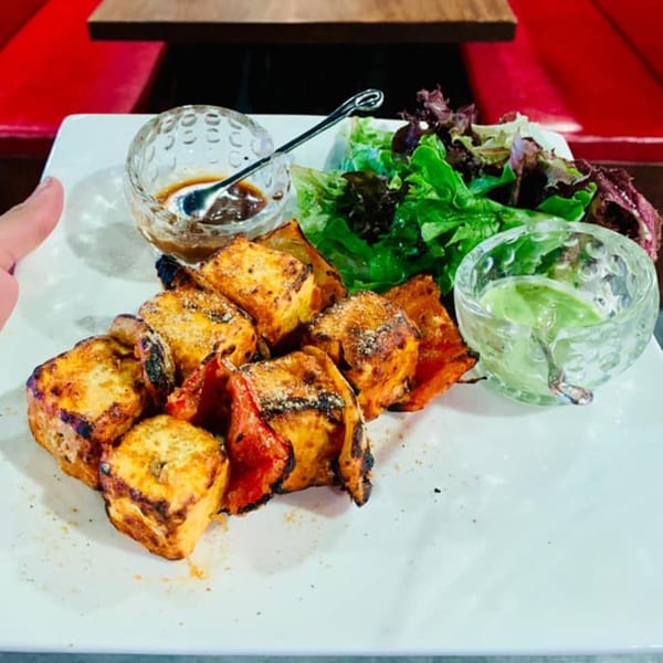 Indian paneer tikka with bell peppers, a side salad, and two dipping sauces on a white plate.
