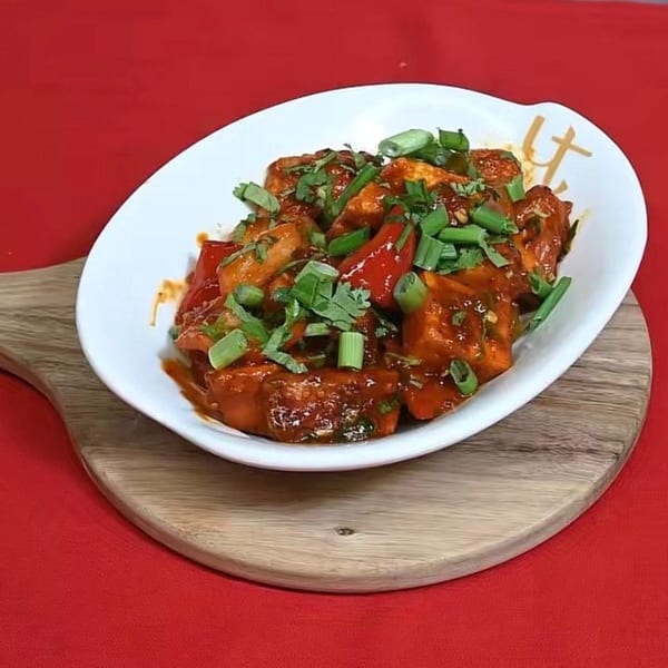 A white bowl of chili paneer with red peppers, green onions, and cilantro on a wooden board.