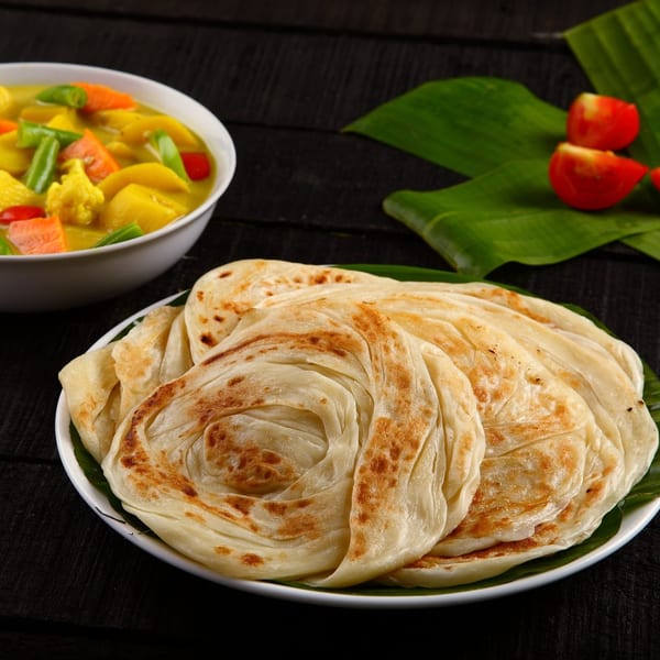 Indian parotta bread on a white plate with a bowl of vegetable curry and banana leaves.
