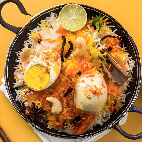 Overhead view of Urban Tandoor's Egg Biryani, garnished with lime, cashews, and spices in a black pan.