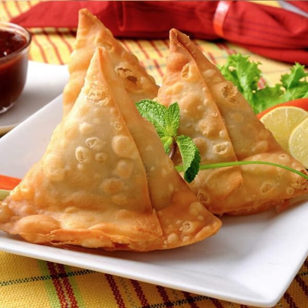 Golden-brown Indian samosas on a white plate with mint, lemon, lettuce, and a side of sauce.