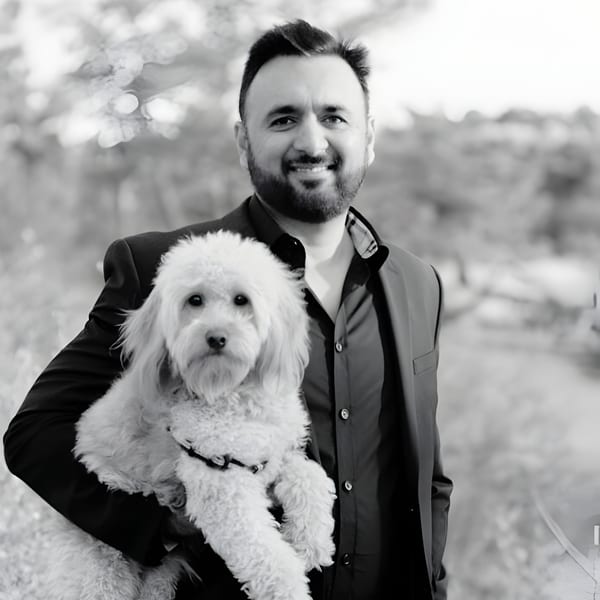 A smiling man in a suit holds a fluffy white dog in a black and white outdoor photo.