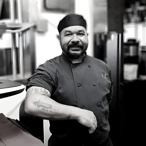 Smiling chef in a black uniform with a "UT" logo and tattooed arm, leaning on a counter.