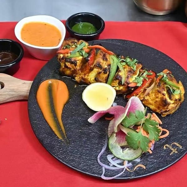 Indian paneer tikka with bell peppers, lemon, salad, and sauces on a black plate.